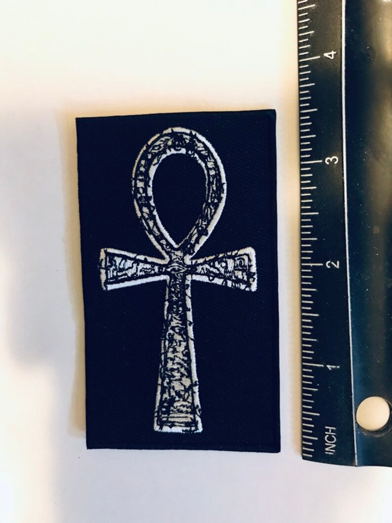 Puede incluir: Un parche de tela negro con un s&iacute;mbolo Ankh bordado en blanco. El parche mide aproximadamente 5 cm de ancho y 7,5 cm de alto.