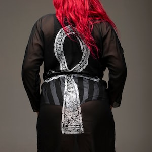 May include: A black sheer robe with a white Ankh symbol printed on the front. The robe has a tie belt and a long, flowing silhouette.