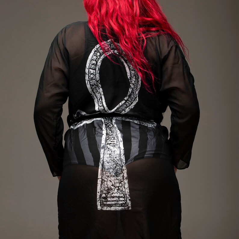 May include: A black sheer robe with a white Ankh symbol printed on the front. The robe has a tie belt and a long, flowing silhouette.