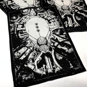 May include: Black and white embroidered patch featuring a detailed illustration of a creature with a skull-like face surrounded by gears and machinery. The patch has a textured fabric surface.