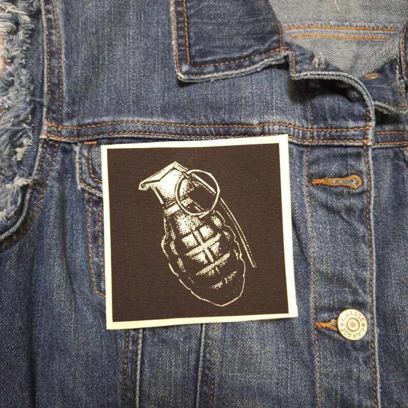 May include: A black and white image of a grenade on a brown patch. The grenade is a common symbol of danger and destruction.