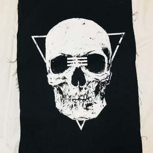 Op de afbeelding: Zwarte stoffen patch met een wit schedelontwerp. De schedel heeft een driehoek erboven en een driehoek eronder. De schedel heeft een witte lijn over de ogen.