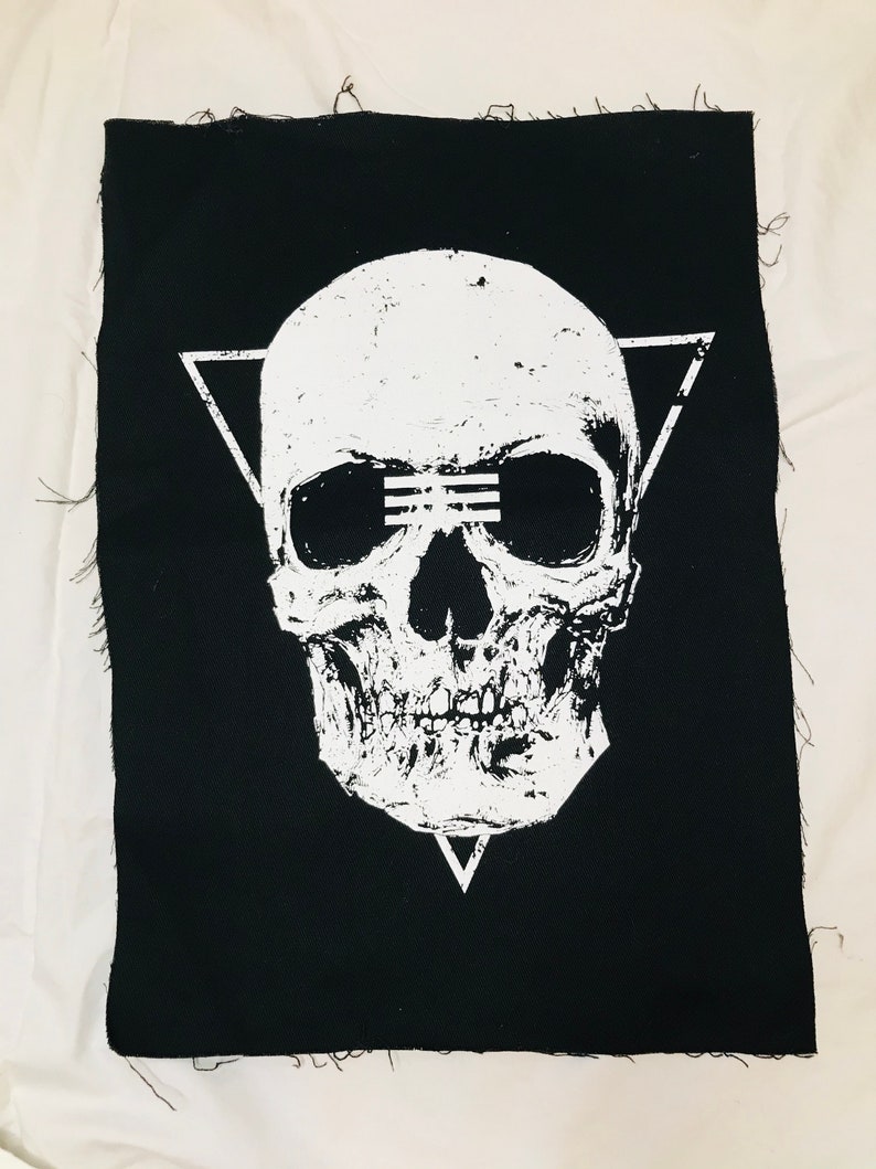 Op de afbeelding: Zwarte stoffen patch met een wit schedelontwerp. De schedel heeft een driehoek erboven en een driehoek eronder. De schedel heeft een witte lijn over de ogen.