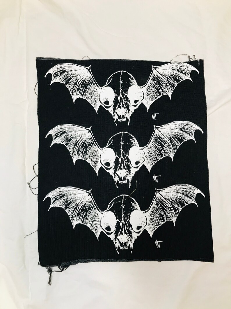 Op de afbeelding: Zwarte stoffen patch met drie witte schedel- en vleermuisontwerpen. Elke schedel heeft grote, ronde ogen en een puntige neus. De vleermuizen hebben grote, uitgespreide vleugels. De tekst "NOT" staat op elke patch.