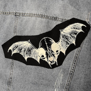 Pode incluir: Remendo preto com uma imagem branca de uma caveira com asas de morcego. A caveira tem duas órbitas oculares grandes e um nariz pontudo.