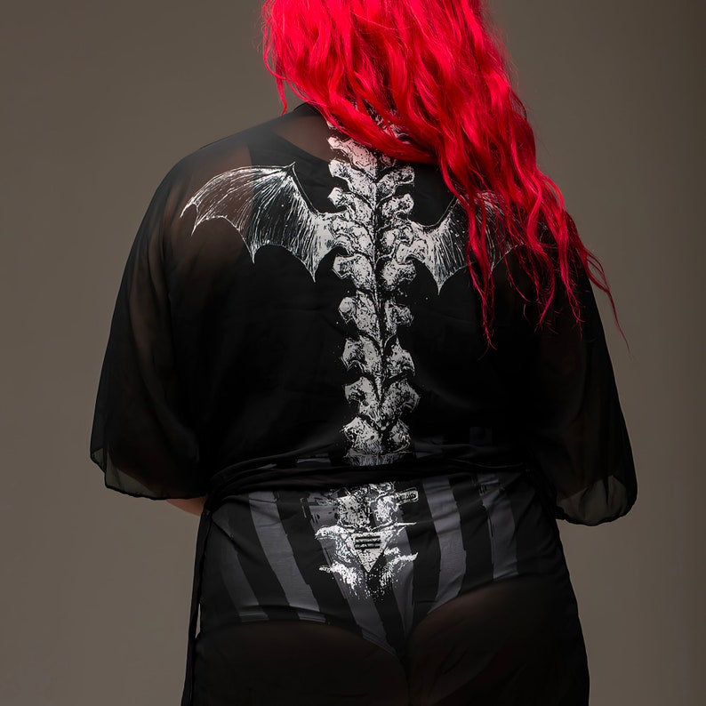 May include: Black sheer kimono with a white spine and bat wing design on the back. The model is wearing black and gray striped underwear with a matching spine design. The model has long, bright red hair.