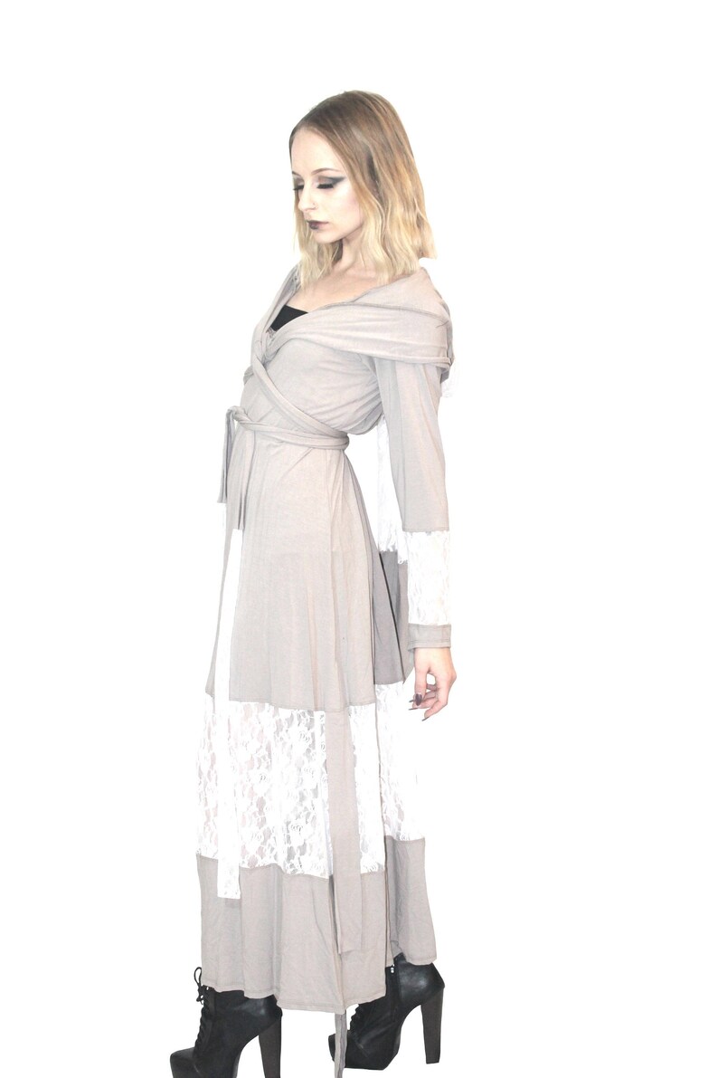May include: A long, gray hooded wrap dress with a white lace trim. The dress has a tie belt and long sleeves. The model is wearing black heeled boots.