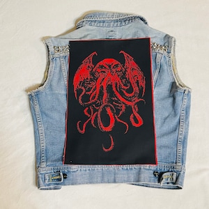 Peut inclure: Un gilet en jean bleu clair avec des clous argentés. Le dos présente un grand patch noir avec une illustration rouge d'une créature avec des tentacules et des ailes. Le gilet a un col classique et des boutons.