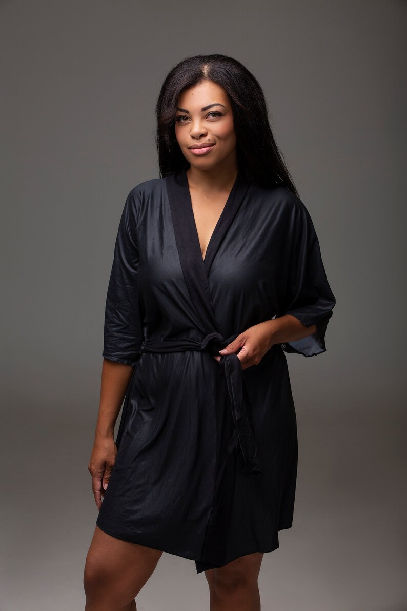 May include: A black satin robe with a shawl collar and a tie belt. The robe is worn by a person with long dark hair.