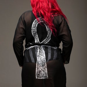 May include: A black sheer robe with a white Ankh symbol printed on the back. The robe has long sleeves and a tie belt. The model has long red hair.