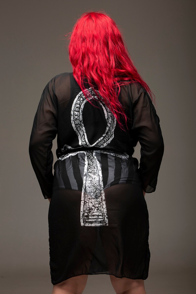 May include: A black sheer robe with a white Ankh symbol printed on the back. The robe has long sleeves and a tie belt. The model has long red hair.