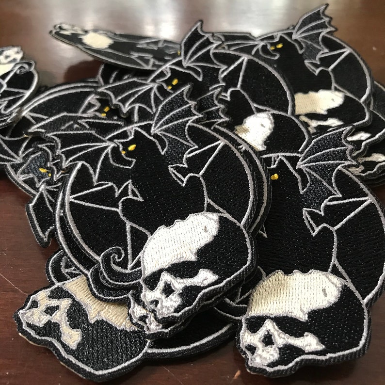 Pode incluir: Uma pilha de patches bordados em preto e branco com um gato preto com asas empoleirado em um cr&acirc;nio. O gato tem olhos amarelos e o cr&acirc;nio tem dentes brancos.