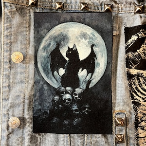 May include: Black cat with bat wings sitting on a pile of skulls in front of a full moon. The cat has yellow eyes and is facing forward. The moon is white and gray and has a dark gray background.