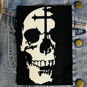 May include: Black and white patch featuring a skull design. The skull is white and has a cross over the top of its head. The cross is also white.