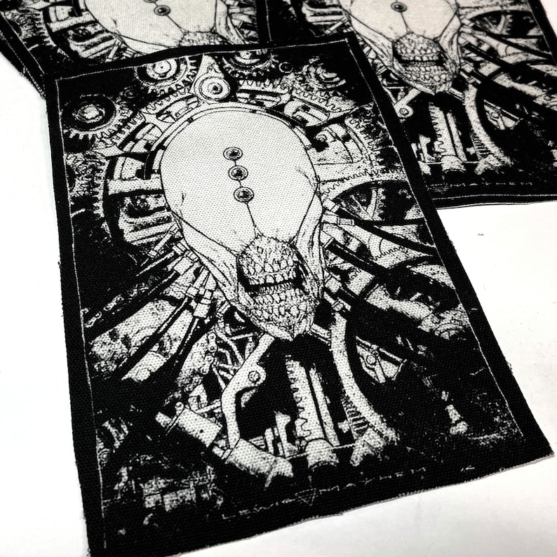 May include: Black and white embroidered patch featuring a detailed illustration of a creature with a skull-like face surrounded by gears and machinery. The patch has a textured fabric surface.
