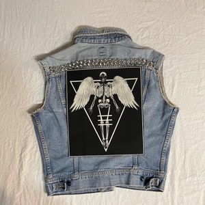 Op de afbeelding: Een lichtblauw denim vest met een zwarte patch met een skelet met vleugels in een driehoek. Het vest is versierd met zilveren studs.