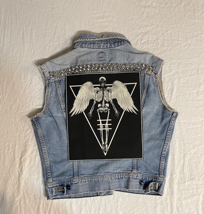 K&ouml;nnte beinhalten: Eine hellblaue Jeansweste mit einem schwarzen Patch, der ein Skelett mit Fl&uuml;geln in einem Dreieck zeigt. Die Weste ist mit silbernen Nieten verziert.