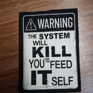 May include: A black and white rectangular patch with the text "WARNING THE SYSTEM WILL KILL YOU TO FEED IT SELF". The patch has a black border and a warning symbol at the top.
