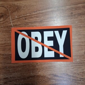 May include: A rectangular patch with the word "OBEY" in large white capital letters on a black background, framed by an orange border. A red diagonal line crosses out the word. The patch is on a wooden surface.