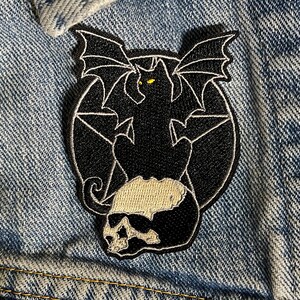 Pode incluir: Etiqueta bordada preta de um gato com asas de morcego sentado em um pentagrama. O gato tem olhos amarelos e uma cauda com forma de caveira.