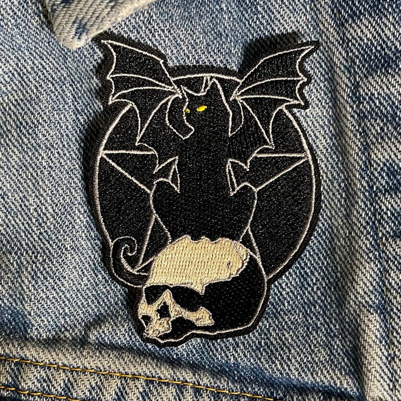 Pode incluir: Etiqueta bordada preta de um gato com asas de morcego sentado em um pentagrama. O gato tem olhos amarelos e uma cauda com forma de caveira.