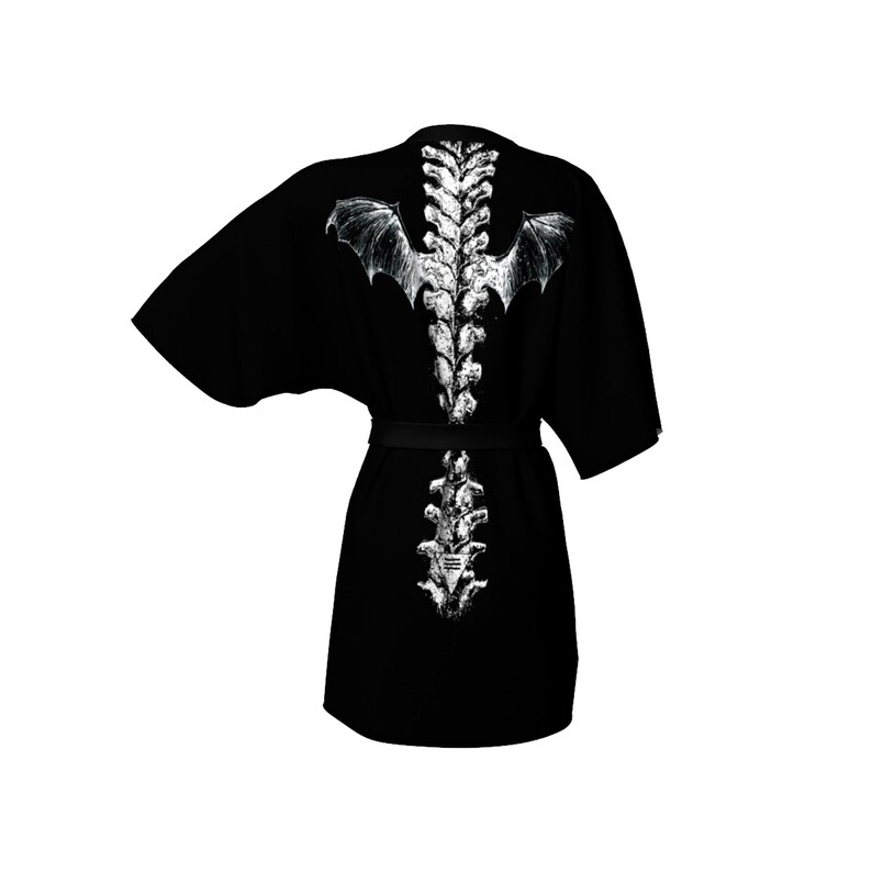 May include: Black kimono-style robe featuring a white spine and bat wing design on the back. The robe has a matching black belt and wide sleeves. The design is a detailed illustration of a spine with bat wings.