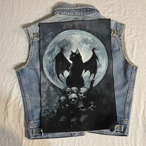 Peut inclure: Un gilet en jean bleu clair avec une peinture en noir et blanc d'un chat noir avec des ailes de chauve-souris devant une pleine lune, perché sur une pile de crânes. Le gilet a des clous argentés sur les épaules.