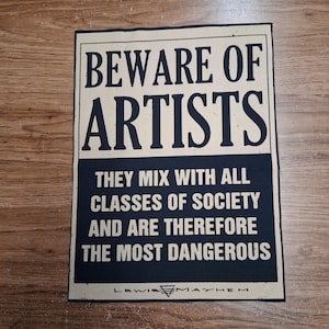 May include: A rectangular sign with the text "BEWARE OF ARTISTS" in large, bold, black letters. Below, smaller text reads "THEY MIX WITH ALL CLASSES OF SOCIETY AND ARE THEREFORE THE MOST DANGEROUS." The sign is on a light beige background.