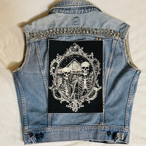 May include: A blue denim vest with silver studs and a black patch featuring a white ornate frame with three skeletons and a house inside.