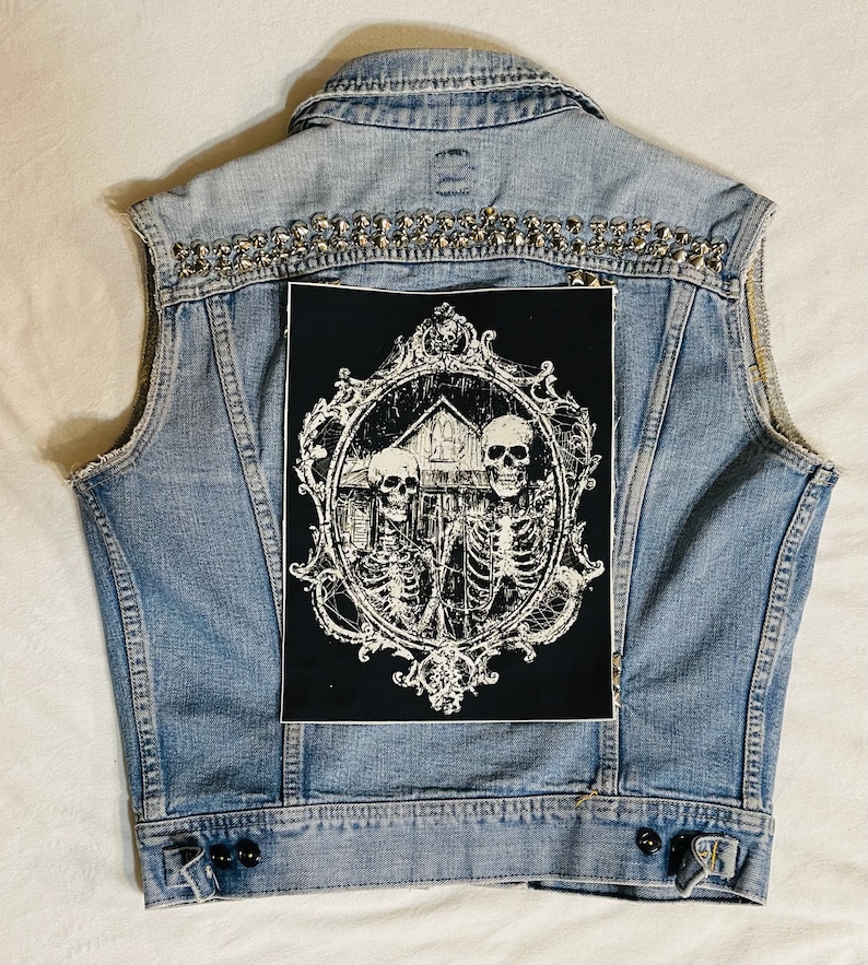 May include: A blue denim vest with silver studs and a black patch featuring a white ornate frame with three skeletons and a house inside.