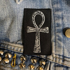 Puede incluir: Un parche termoadhesivo negro con un s&iacute;mbolo Ankh bordado en blanco. El Ankh es un s&iacute;mbolo jerogl&iacute;fico egipcio antiguo que representa la vida.