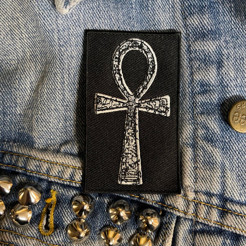 Puede incluir: Un parche termoadhesivo negro con un s&iacute;mbolo Ankh bordado en blanco. El Ankh es un s&iacute;mbolo jerogl&iacute;fico egipcio antiguo que representa la vida.