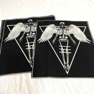 K&ouml;nnte beinhalten: Zwei schwarze Stoffpatches mit einem wei&szlig;en Skelett mit Fl&uuml;geln in einem wei&szlig;en Dreieck. Das Skelett steht mit ausgestreckten Armen. Die Patches liegen auf einer wei&szlig;en Oberfl&auml;che.