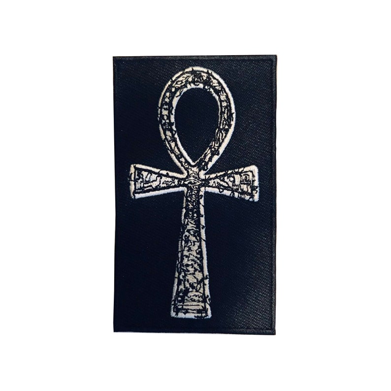 Puede incluir: Un parche de tela negro con un s&iacute;mbolo Ankh bordado en blanco.