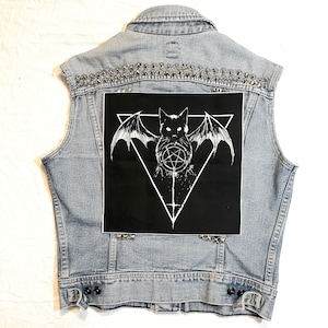 Peut inclure: Un gilet en jean bleu clair avec des clous argentés et un patch noir représentant une illustration blanche d'un chat avec des ailes de chauve-souris et un pentagramme.