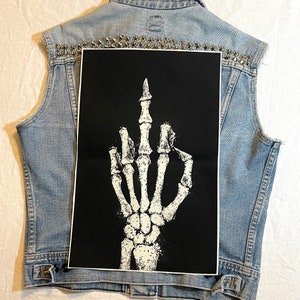 May include: A black and white patch featuring a skeleton hand giving the middle finger. The patch is attached to a denim vest with silver studs.