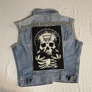 Op de afbeelding: Een blauwe denimvest met zilveren studs en een zwarte patch met een witte schedel met een gasmasker en een geometrisch ontwerp.