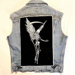 May include: A light blue denim vest with a black and white graphic of a skeleton with wings and a scythe. The vest has silver studs along the top and black buttons.