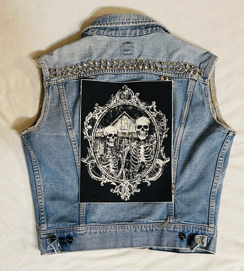 May include: A blue denim vest with silver studs and a black patch featuring a white ornate frame with three skeletons and a house inside.