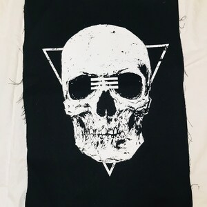Op de afbeelding: Zwarte stoffen patch met een wit schedelontwerp. De schedel heeft een driehoek erboven en een driehoek eronder. De schedel heeft een witte lijn over de ogen.