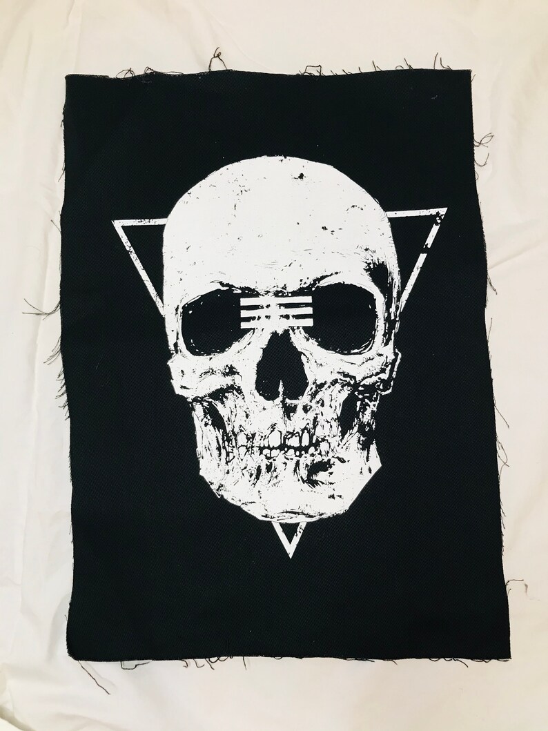 Op de afbeelding: Zwarte stoffen patch met een wit schedelontwerp. De schedel heeft een driehoek erboven en een driehoek eronder. De schedel heeft een witte lijn over de ogen.