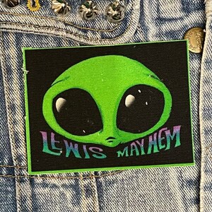 Pode incluir: Um remendo preto com um rosto de alienígena verde e o texto "Lewis Mayhem" em um degradê arco-íris.