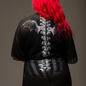 May include: Black sheer kimono with a white spine and bat wing design on the back. The model is wearing black and gray striped underwear with a matching spine design. The model has long, bright red hair.