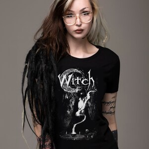 May include: A black t-shirt with a white graphic of a crescent moon and the word "Witch" in a gothic font. The graphic also features a stylized figure with a long, flowing tail.