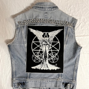 May include: A denim vest with silver studs and a black patch featuring a white illustration of an angel with wings, holding a sword, standing in a circle with a star inside. The angel is surrounded by a circle with a star inside.