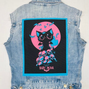 Peut inclure: Patch thermocollante noir et bleu avec un chat noir de dessin animé aux yeux roses et aux ailes debout sur une pile de crânes roses et blancs. Le patch a une lune rose et bleue en arrière-plan avec des chauves-souris qui volent autour. Le texte "Lewis Mayhem Presents Baby Mina" est en bas du patch.
