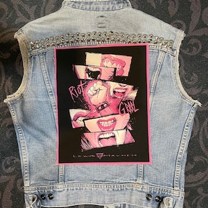 Op de afbeelding: Een lichtblauw denim vest met zilveren studs en een zwart en roze patch met de tekst "Riot Grrrl" met een roze driehoek en de tekst "Lewis Mayhem" onderaan.