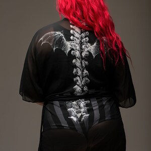 May include: Black sheer kimono-style top and shorts set with a white skeletal spine and bat wing design on the back. The shorts have a striped pattern. The model has bright red hair.