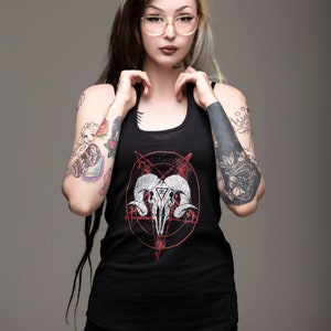 May include: A woman with black and blonde hair wearing a black tank top with a red and white graphic of a skull with horns and a pentagram. She has several tattoos on her arms and legs.