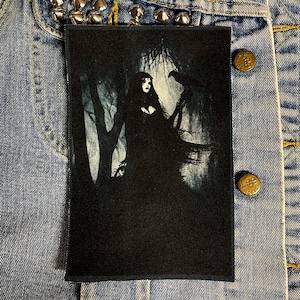 May include: Black and white embroidered patch featuring a woman with long dark hair standing in a forest with a raven perched on her arm. The patch is on a denim background.